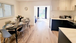 A modern kitchen with white cabinets, a dining area with a table set for four, and a view into a bright dining room. - Exclusive Palm House,  Nature, Amenities, Garden (Addlestone)