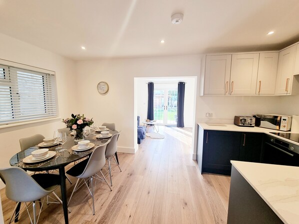 A modern kitchen with white cabinets, a dining area with a table set for four, and a view into a bright dining room. - Exclusive Palm House, Nature, Amenities, Garden (Addlestone)