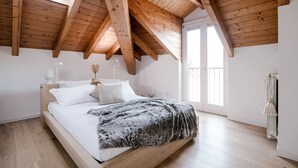 A bedroom with a sloped wooden ceiling, a bed with white bedding, a fluffy throw blanket, and a large window providing natural light. - MyHomeInComo - Modern Luxury Attic (Como)