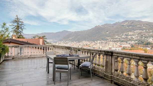 A terrace with a table and chairs, overlooking a cityscape with mountains in the background. - MyHomeInComo - Modern Luxury Attic (Como)
