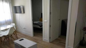 Ein Zimmer mit Glastisch, weißen Stühlen, einem weißen Schrank, einem Bett mit weißer und grauer Bettdecke und einem an der Wand befestigten Fernseher.