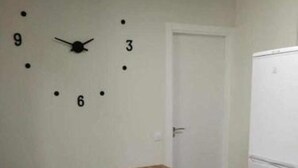 Ein Zimmer mit einer Wanduhr, einem Holztisch, einem weißen Kühlschrank und einer Tür.