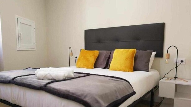 Ein ordentlich bezogenes Bett mit einer grau-weißen Decke, zwei gelben Kissen und einem schwarzen Kopfteil. Ein Nachttisch mit Lampe und Steckdose ist ebenfalls vorhanden.