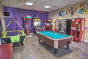 Ein Billardtisch, Spielautomaten und eine Coca-Cola-Getränkeautomat in einem Raum mit violetten Wänden und Cartoon-Verzierungen.