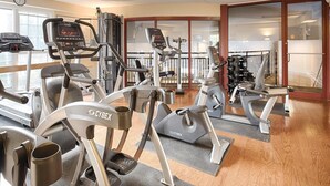 A gym with various cardio machines, including treadmills and elliptical trainers, arranged in a spacious layout with large windows and a wooden floor.