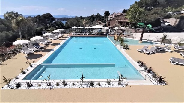 A rectangular swimming pool with lounge chairs and umbrellas around it, set against a backdrop of trees and a clear sky.