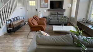Un salon spacieux avec un escalier, un canapé gris, un fauteuil marron, une télévision et une plante.