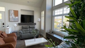 Un salon avec un canapé gris, un canapé marron, une table basse blanche et un téléviseur fixé au mur. De grandes fenêtres sont équipées de stores, et on y trouve une plante ainsi qu’un tableau accroché au mur.