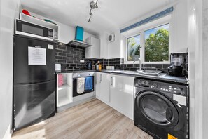 Een moderne keuken met een zwarte koelkast, magnetron en wasmachine. Een raam zorgt voor daglicht en uitzicht op bomen.