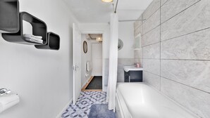 Een moderne badkamer met een witte badkuip, een wastafel en een douchegedeelte. Er zijn planken aan de muur en een vloerkleed met een patroon op de vloer.