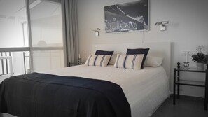 A hotel room with a large bed, a cityscape painting, a nightstand, and a view through a large window.
