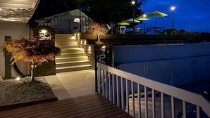 Un espace extérieur aménagé pour la nuit, avec un escalier menant à un espace de repos abrité, entouré d’arbres et éclairé par des lampadaires.