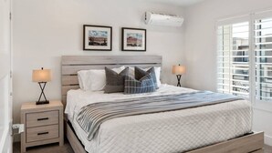 A bedroom with a bed, two bedside tables, a ceiling fan, an air conditioning unit, and two framed pictures on the wall.