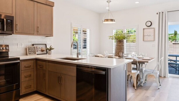A modern kitchen with wooden cabinets, a dishwasher, and a large island. There is a dining area with a table and chairs, and a view of the outdoors through large windows.