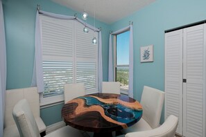 A room with a round wooden table, white chairs, a window with blinds, and a ceiling light fixture.