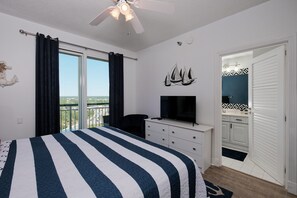 A bedroom with a bed, a TV, a ceiling fan, a window with curtains, a desk, and a bathroom visible through an open door.