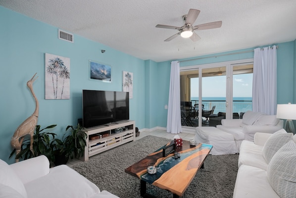 A living room with a TV, white furniture, a wooden coffee table, and a view of the ocean.