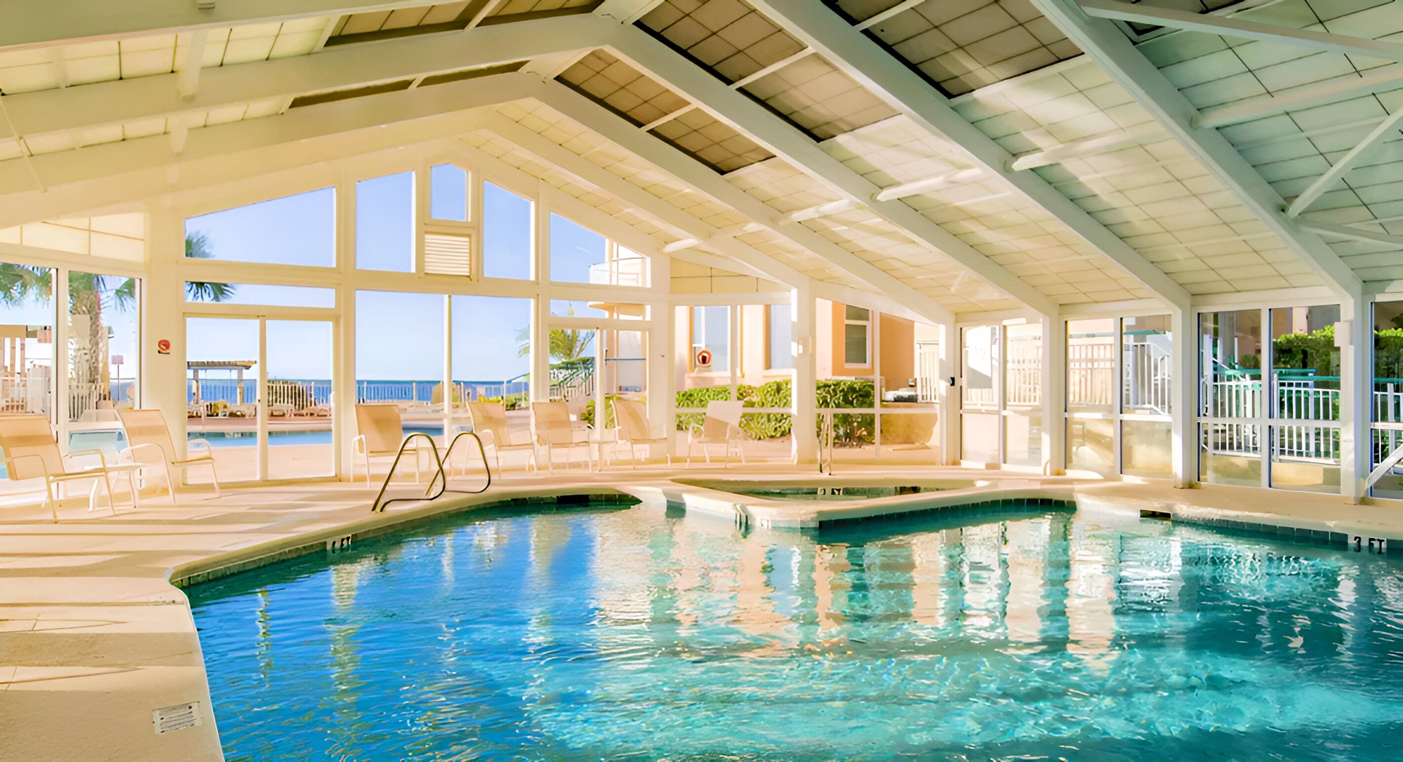An indoor swimming pool with clear glass walls, lounge chairs, and a view of the ocean.