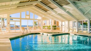 Uma piscina interior com paredes de vidro transparente, espreguiçadeiras e vista para o oceano.