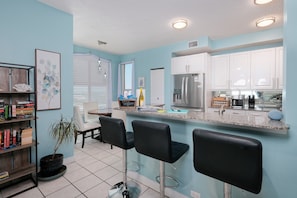A kitchen with white cabinets, a stainless steel refrigerator, a bar area with black stools, and a potted plant.