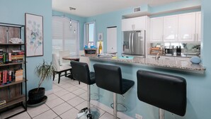 A kitchen with white cabinets, a stainless steel refrigerator, a bar area with black stools, and a potted plant.
