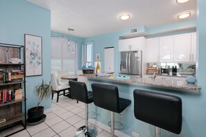 A kitchen with white cabinets, a stainless steel refrigerator, a bar area with black stools, and a potted plant.