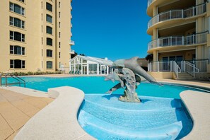 A swimming pool with a dolphin sculpture, a building with balconies, and a clear blue sky.