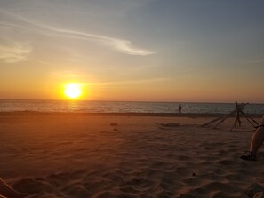 A beach at sunset with a clear sky, calm sea, and a few people and a wooden structure on the sand.