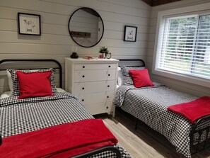 A room with two beds, each with a checkered bedspread and red pillows. There is a dresser with a mirror above it, and two framed pictures on the wall.