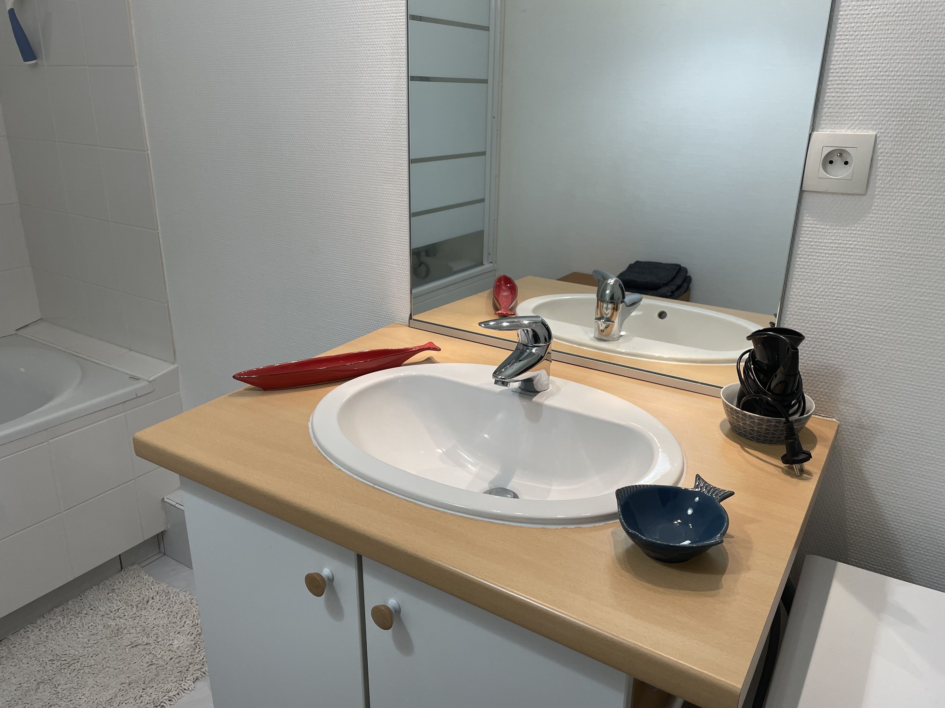 Une salle de bain avec un lavabo blanc, un miroir, un porte-savon rouge, une bouilloire électrique noire et un bol bleu.