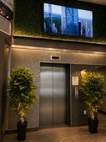 Egy digitális kijelzővel ellátott lift, melynek mindkét oldalán virágtartóban lévő növények állnak, és a felette lévő zöld falra egy televízió van felszerelve.