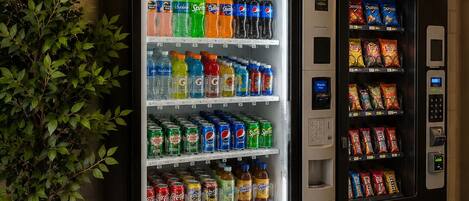 A vending machine with a variety of beverages and snacks, a security camera, and a potted plant.