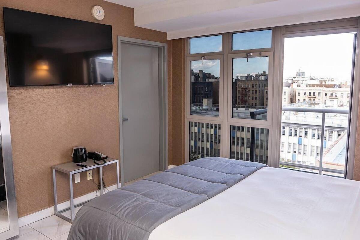 A hotel room with a bed, a TV mounted on the wall, a small table with a phone and a coffee maker, and large windows offering a city view.
