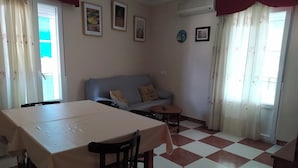 Un salon comprenant un canapé, une table à manger et une petite table ronde. Il y a des rideaux aux fenêtres et quelques tableaux encadrés au mur.