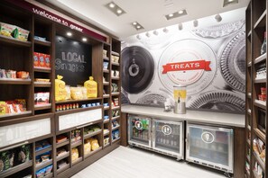 A snack and treat section with shelves stocked with various snacks, a chalkboard sign, and beverage dispensers. - Hampton Inn Greer Greenville, SC (Greer)