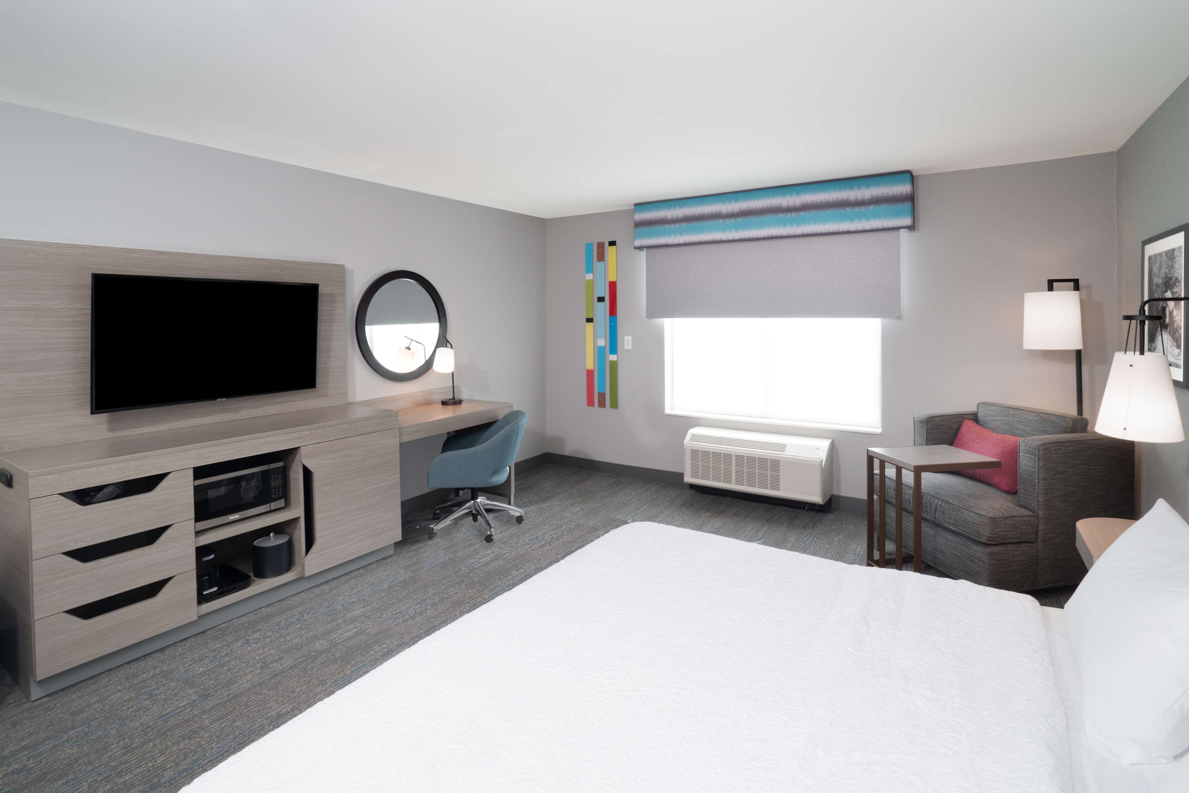 A hotel room with a large bed, a desk with a chair, a flat-screen TV, a round mirror, a window with a view, and a small table with a lamp.