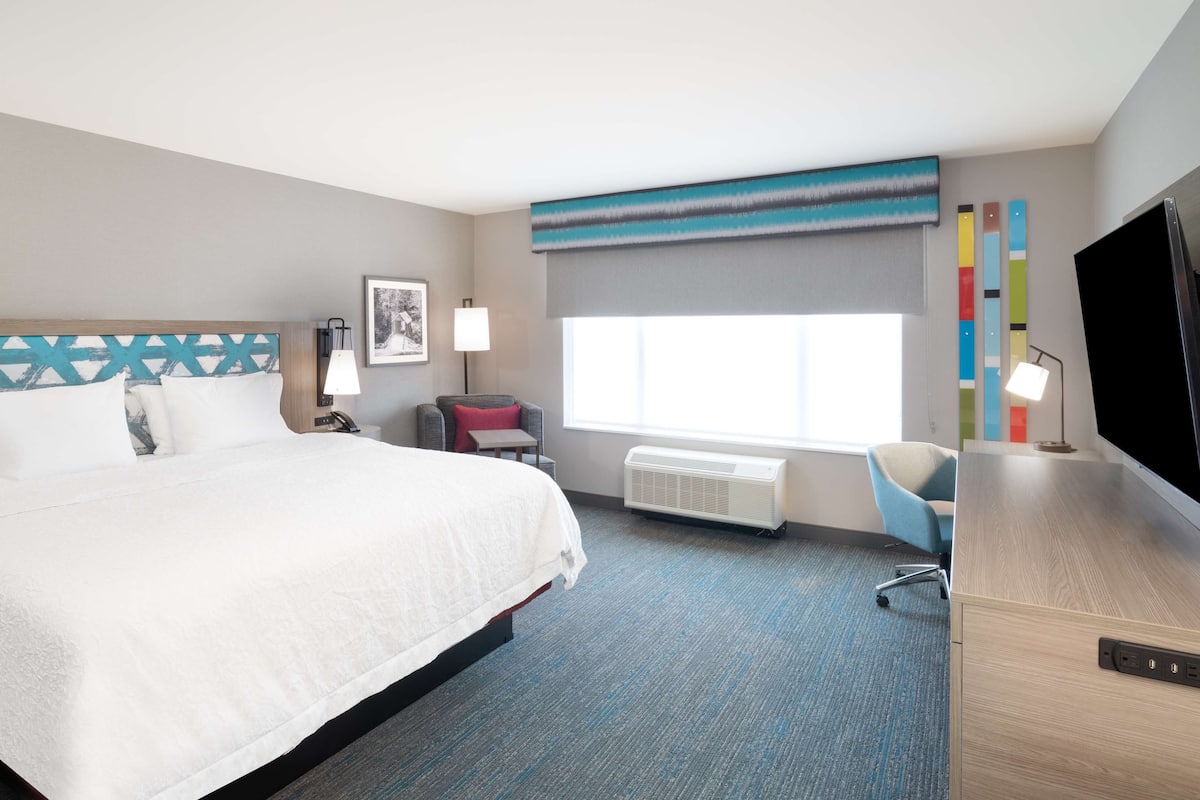 Habitación de hotel con cama, escritorio, silla, televisor, lámpara y aire acondicionado.