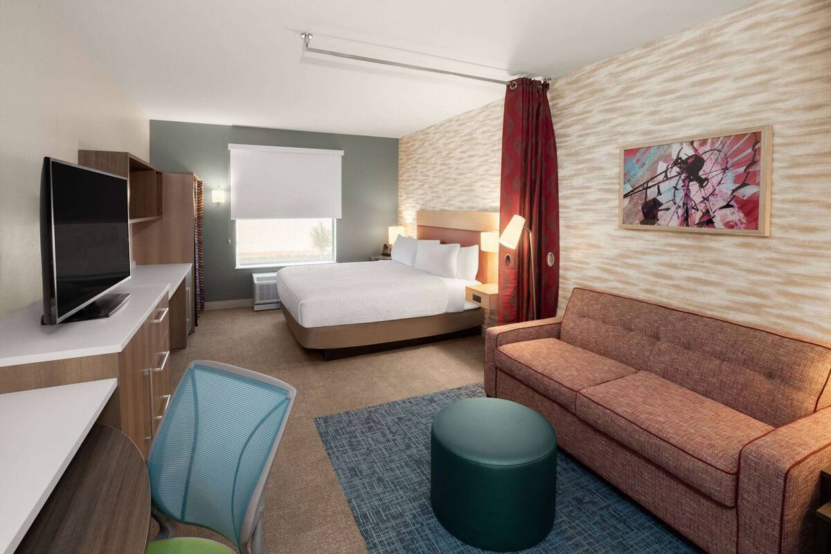 A hotel room with a bed, a desk with a TV, a chair, a sofa, and a painting on the wall.