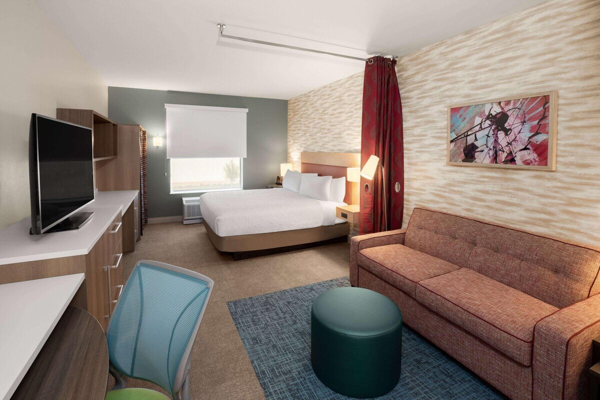 A hotel room with a bed, a desk with a TV, a sofa, a chair, and a painting on the wall.