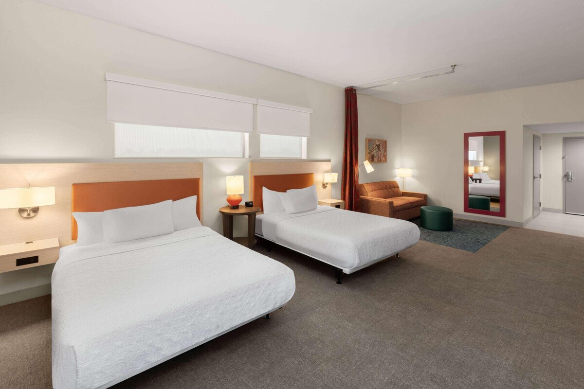 A hotel room with two beds, a chair, a small table, and a mirror.