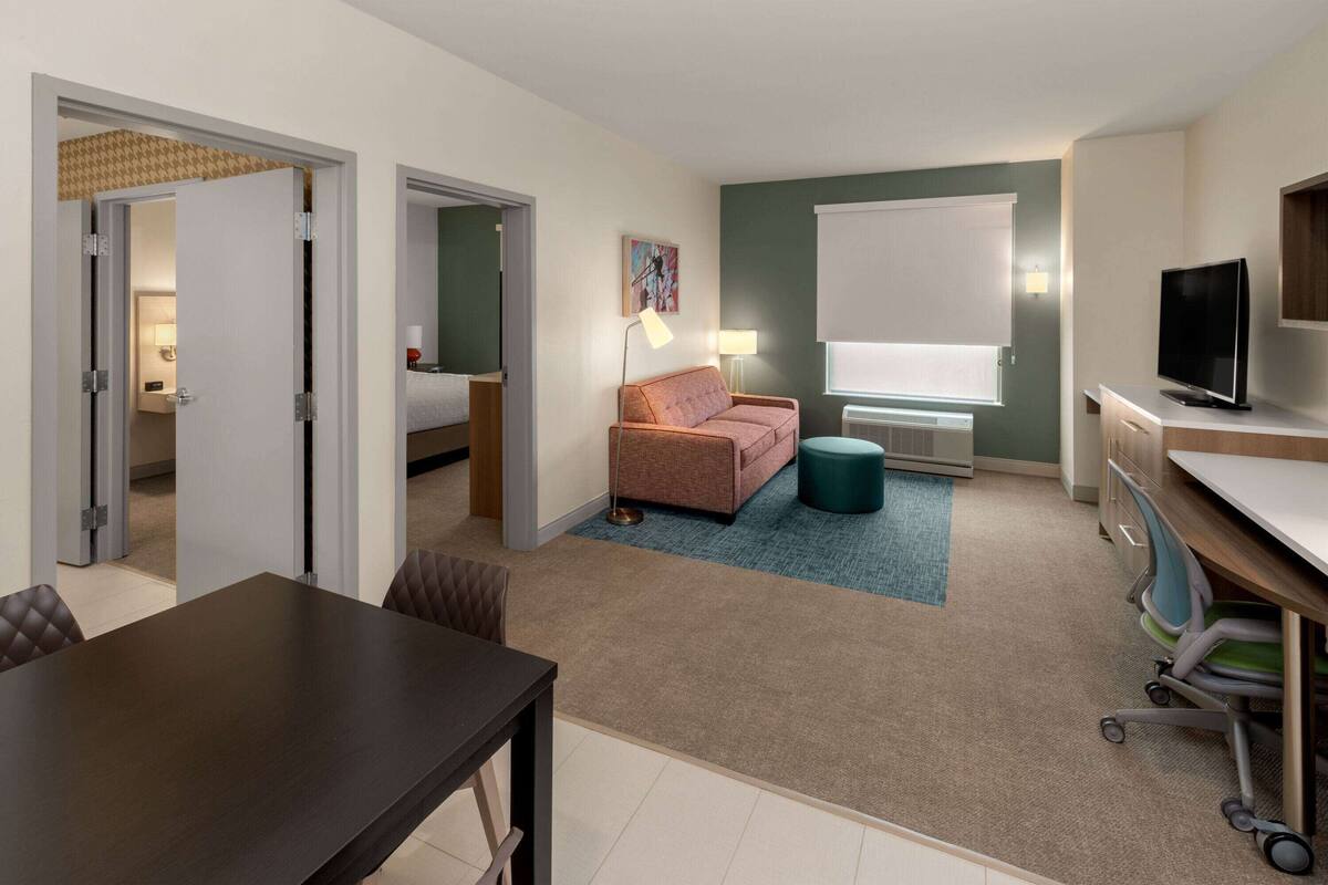 A hotel room with a dining table, a sofa, a TV, and a window with blinds.