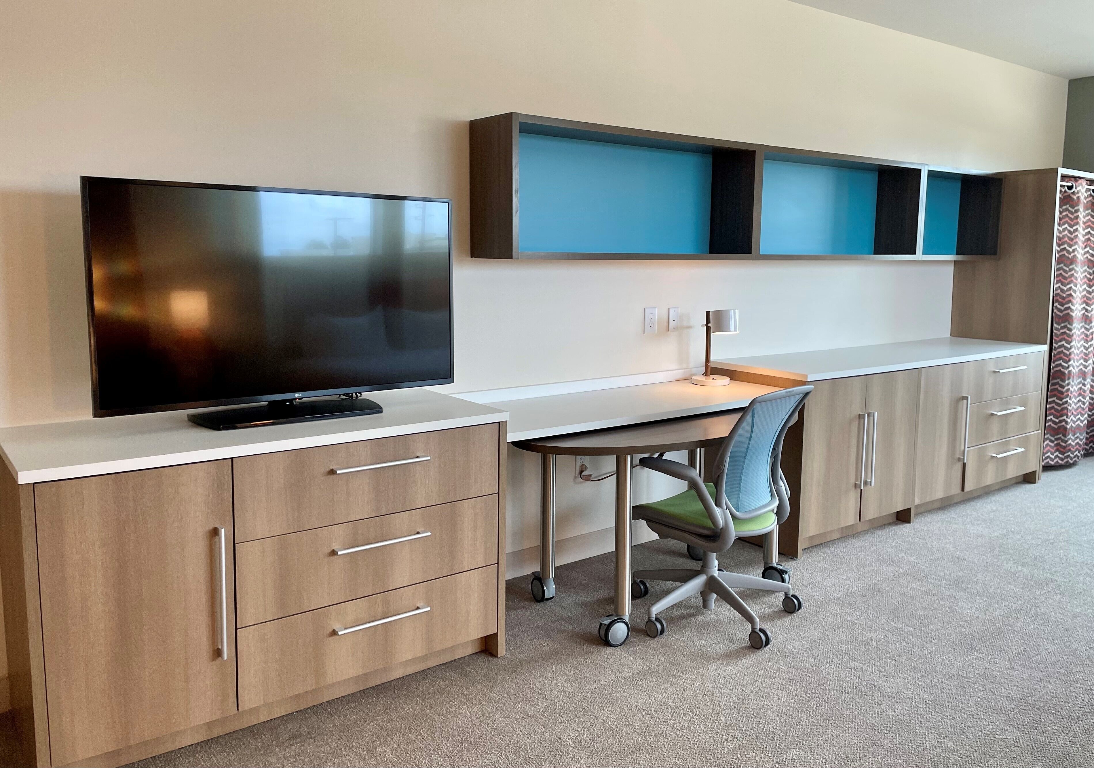 A modern office space with a flat-screen TV, a desk with a chair, and built-in shelving.