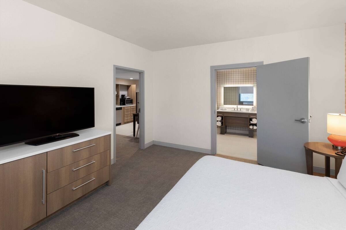 A hotel room with a flat-screen TV on a wooden dresser, a bed with white bedding, a small table with a lamp, and a bathroom visible through an open door.