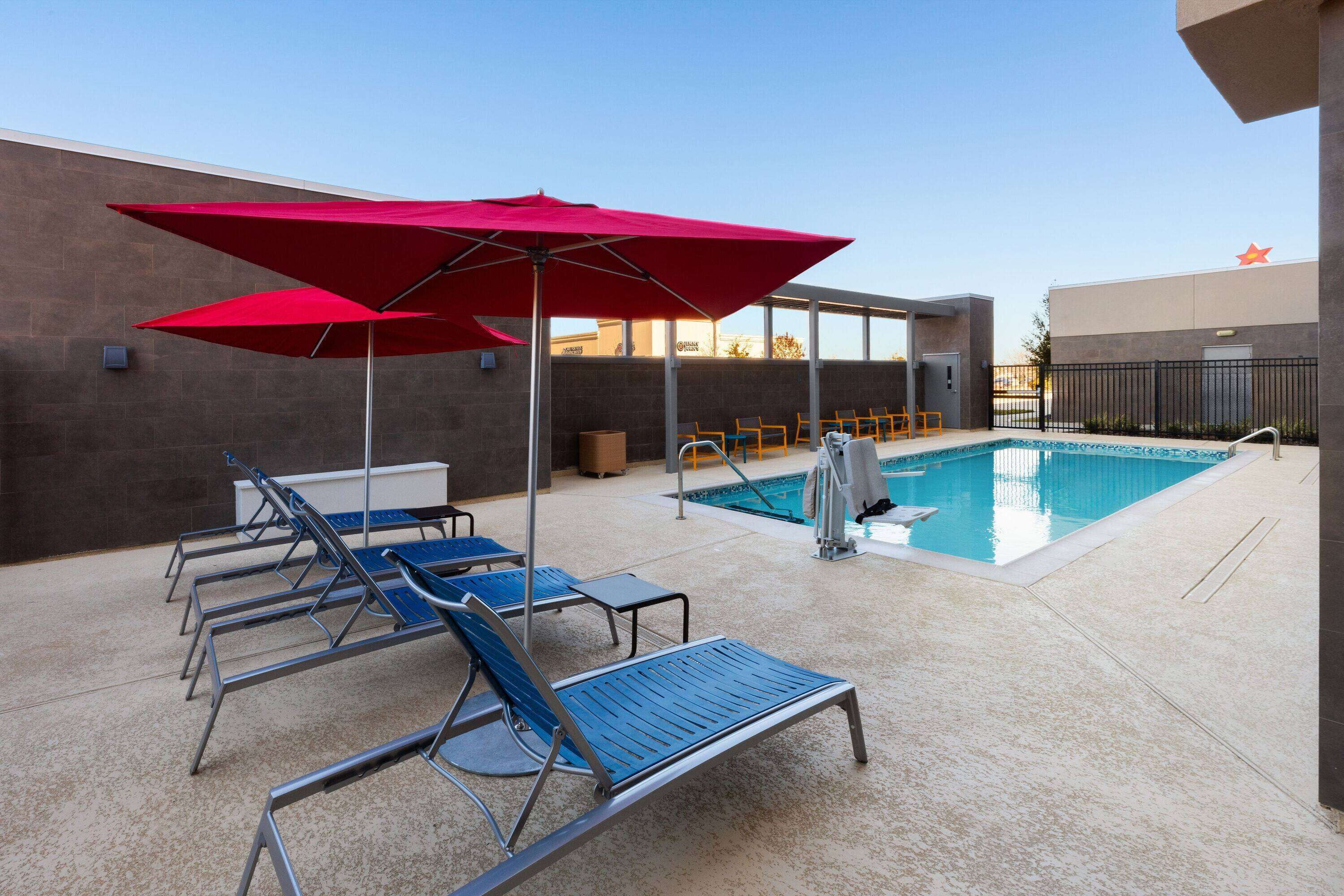 A pool area with blue lounge chairs and red umbrellas, a swimming pool, and a building with a star emblem.