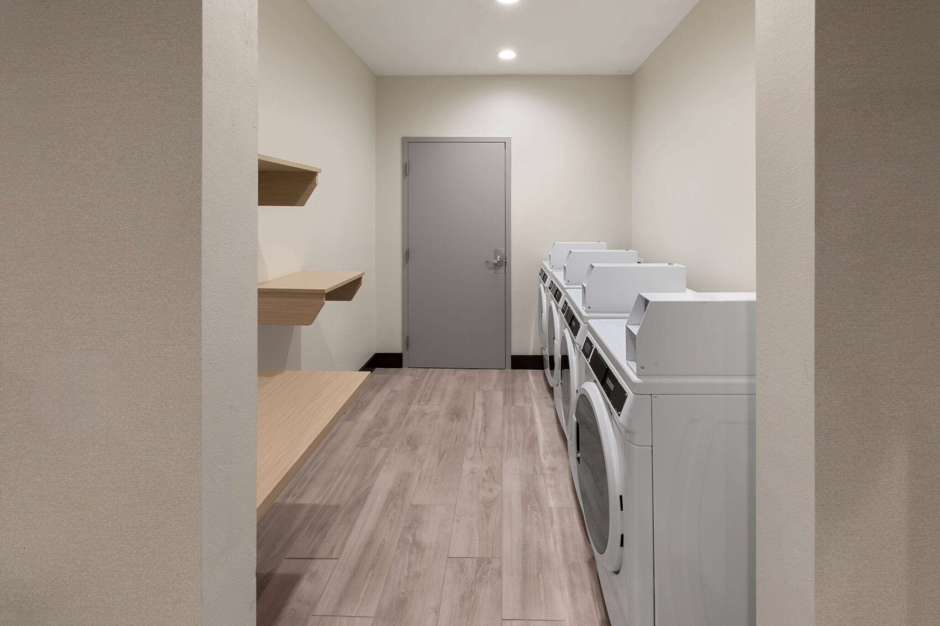 A laundry room with a door, wooden shelves, and a row of washing machines.