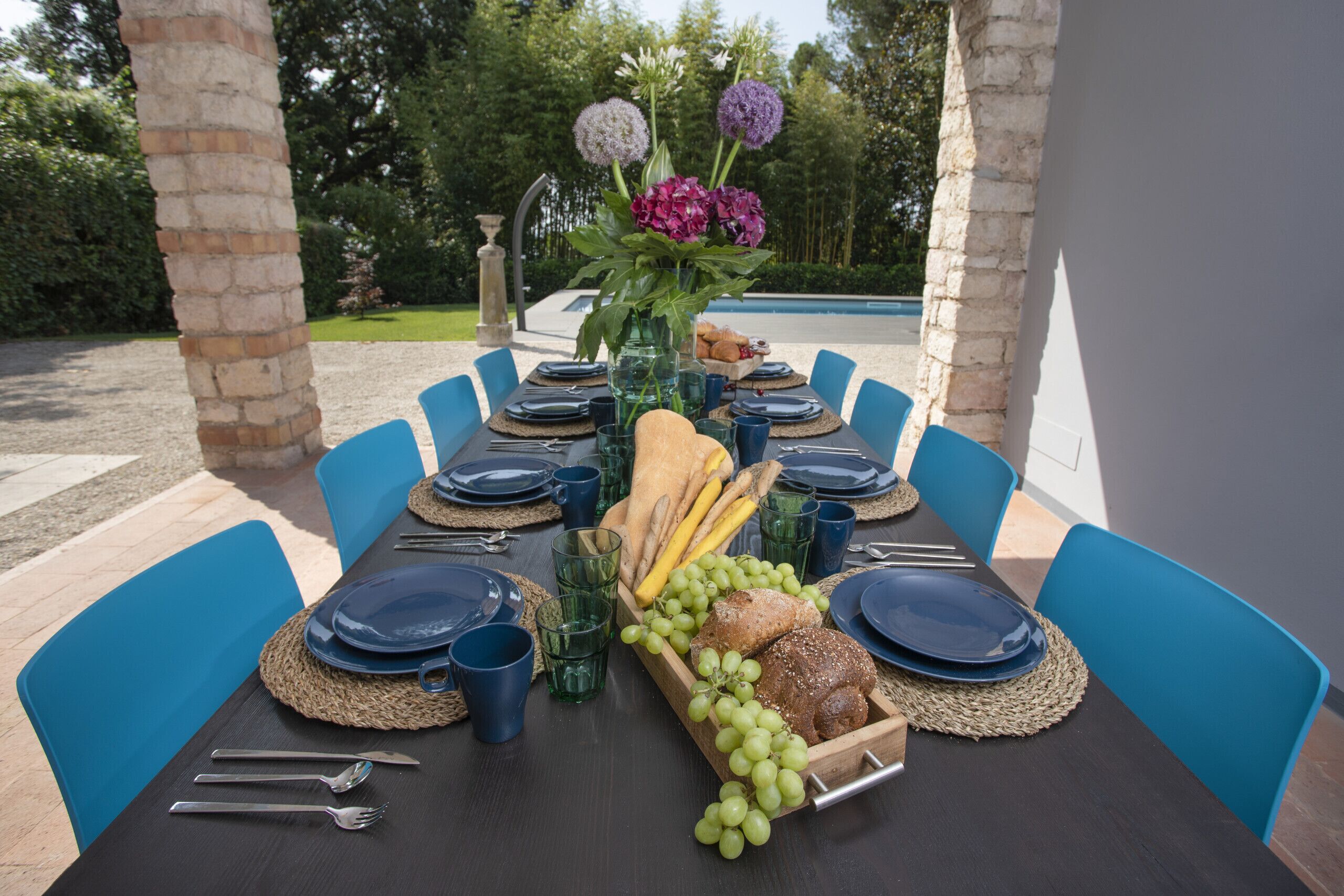 Une table à manger dressée pour six personnes, avec des chaises bleues, entourée d’une piscine et de verdure.