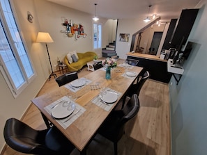 A dining area with a wooden table set for four, black chairs, a yellow sofa, and a modern kitchen in the background. - Bleautiful Eugénie charming townhouse (Fontainebleau)