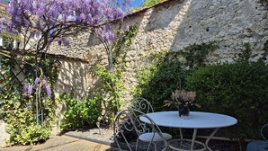 A courtyard with a white table and chairs, surrounded by greenery and purple flowers.
