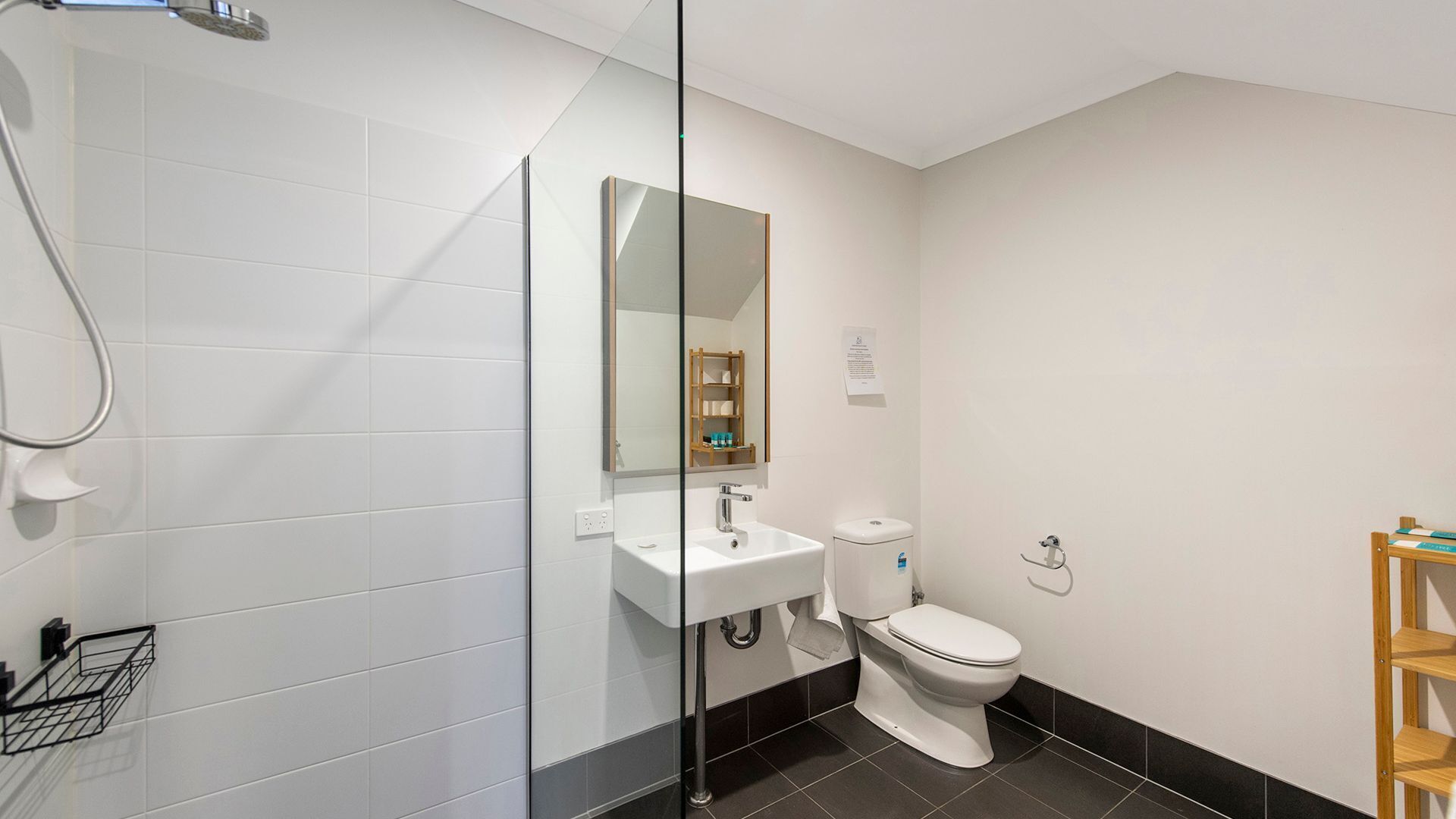A modern bathroom with white tiles, a glass shower enclosure, a white sink, a toilet, and a wooden shelf.