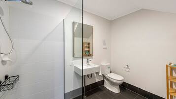 A modern bathroom with white tiles, a glass shower enclosure, a white sink, a toilet, and a wooden shelf.
